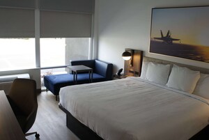 Quarto de hotel com uma cama grande, uma escrivaninha, uma cadeira, uma mesinha e uma janela com vista para um avião no chão.