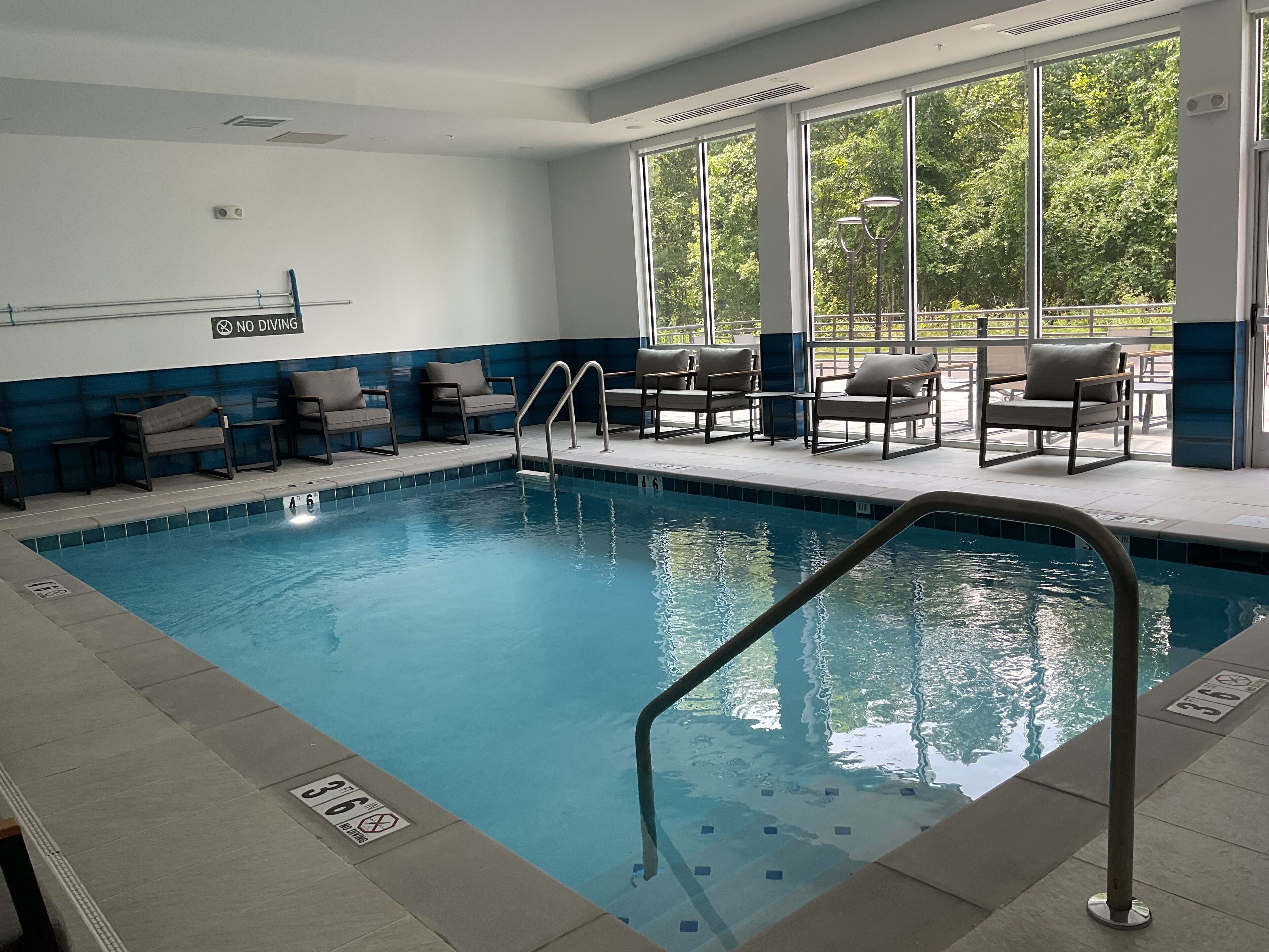 An indoor swimming pool with a 'No Diving' sign, lounge chairs, and large windows offering a view of greenery.