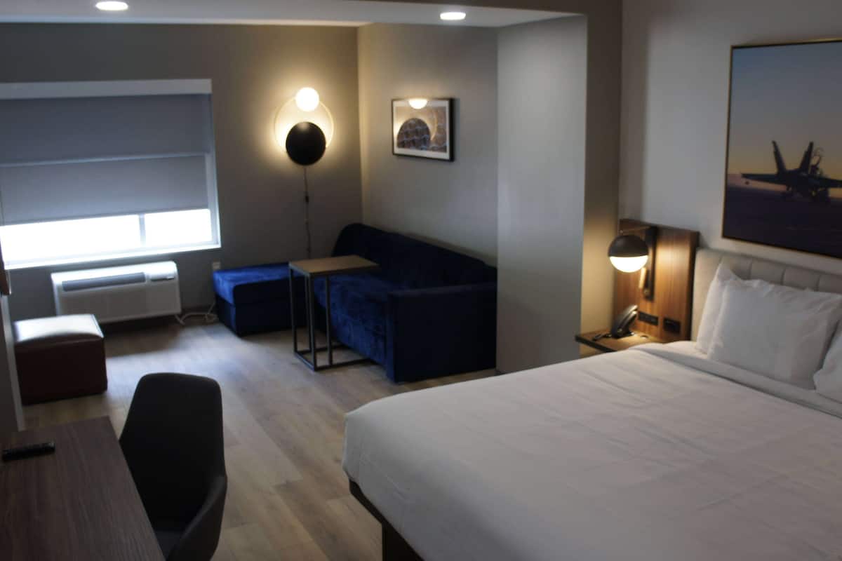 Ein Hotelzimmer mit Bett, Sofa, Schreibtisch und Stuhl. Es gibt ein Fenster mit Jalousien, eine Wandlampe und ein gerahmtes Bild an der Wand.
