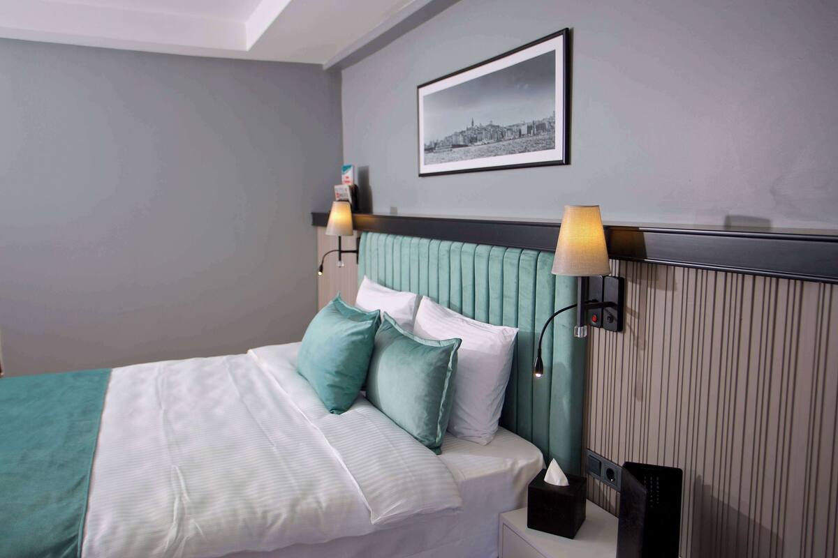Una cama bien tendida con una colcha azul aguamarina y blanca, dos almohadas y una mesita de noche con una lámpara y una caja de pañuelos.