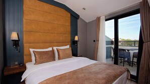 Una habitación de hotel con una cama grande, dos lámparas de noche, vistas al exterior a través de un ventanal corredero y un balcón con sillas.