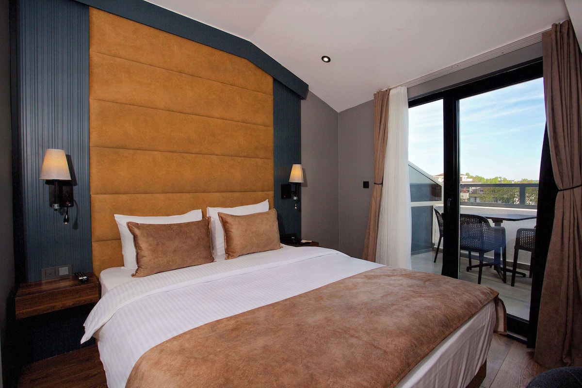 Una habitación de hotel con una cama grande, dos lámparas de noche, vistas al exterior a través de un ventanal corredero y un balcón con sillas.