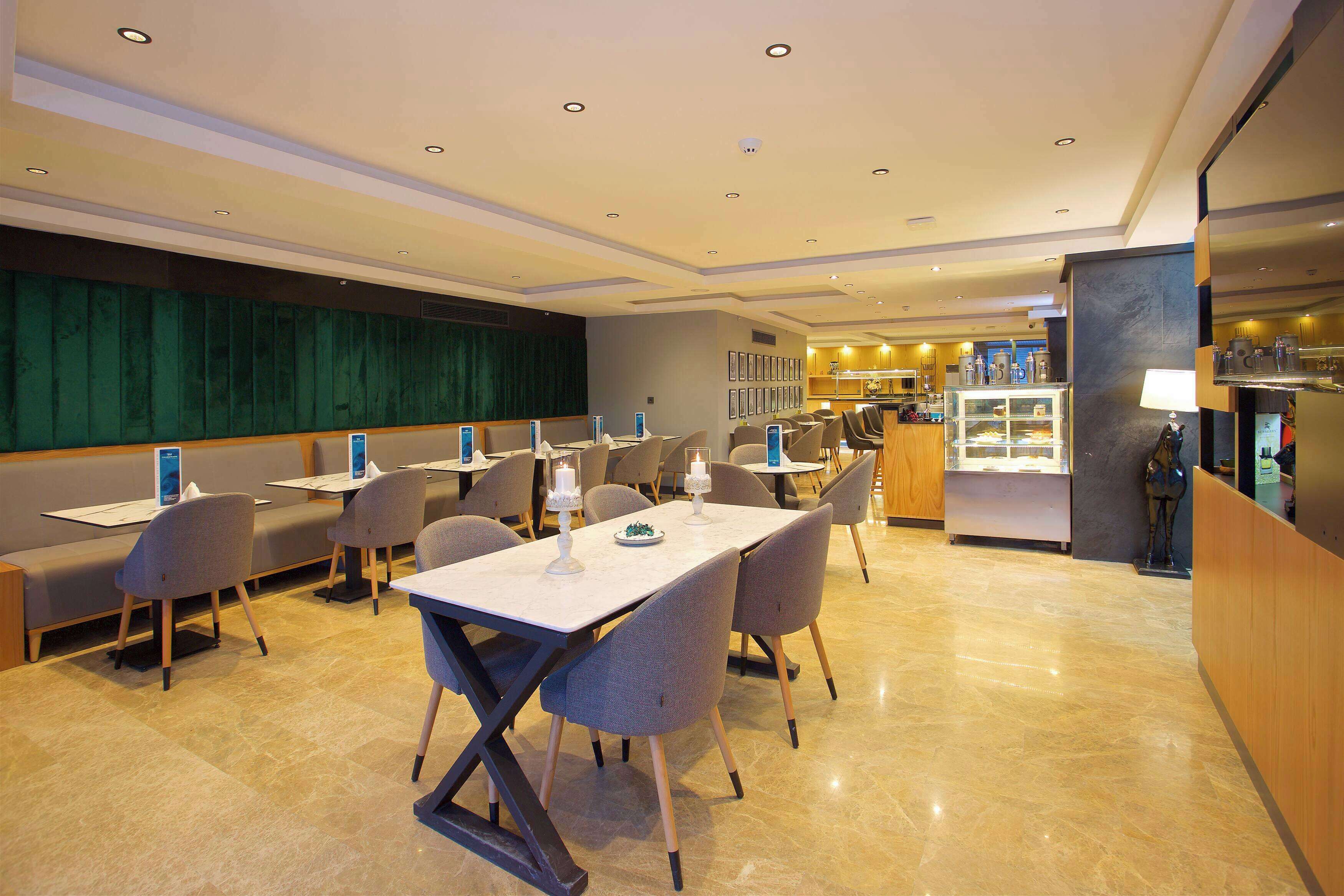 Ein Hotel-Speisesaal mit einem langen Tisch, mehreren Stühlen, einer Theke mit einer Auslage und einer Sitzecke mit grüner Wand.