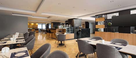 A modern restaurant with marble-topped tables, grey upholstered chairs, and a central bar area.