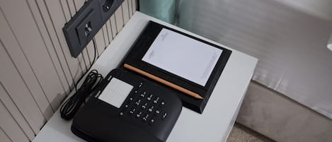 A hotel room with a white bedside table, a black telephone, a notepad, and a pen.