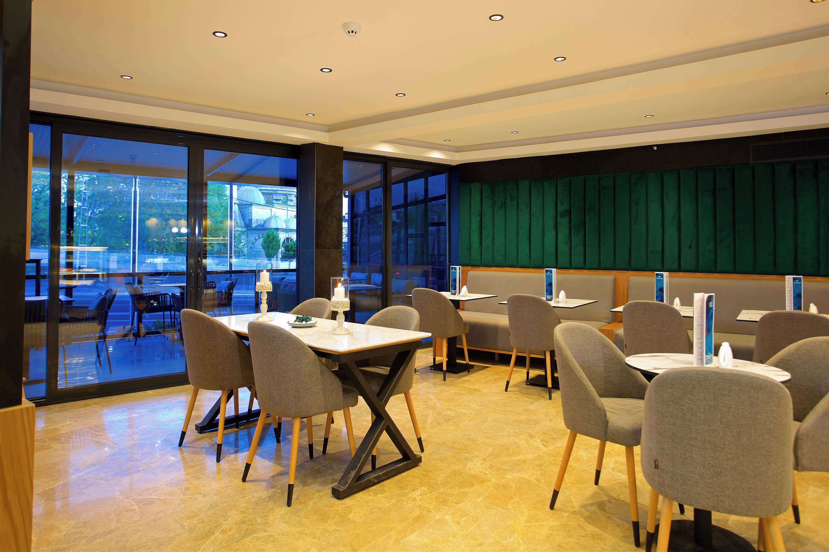 A modern dining area with tables and chairs, large windows, and a green wall.