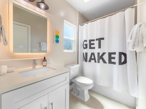 Um banheiro com pia branca, espelho e vaso sanitário. Cortina de chuveiro com a inscrição 'GET NAKED' é visível.