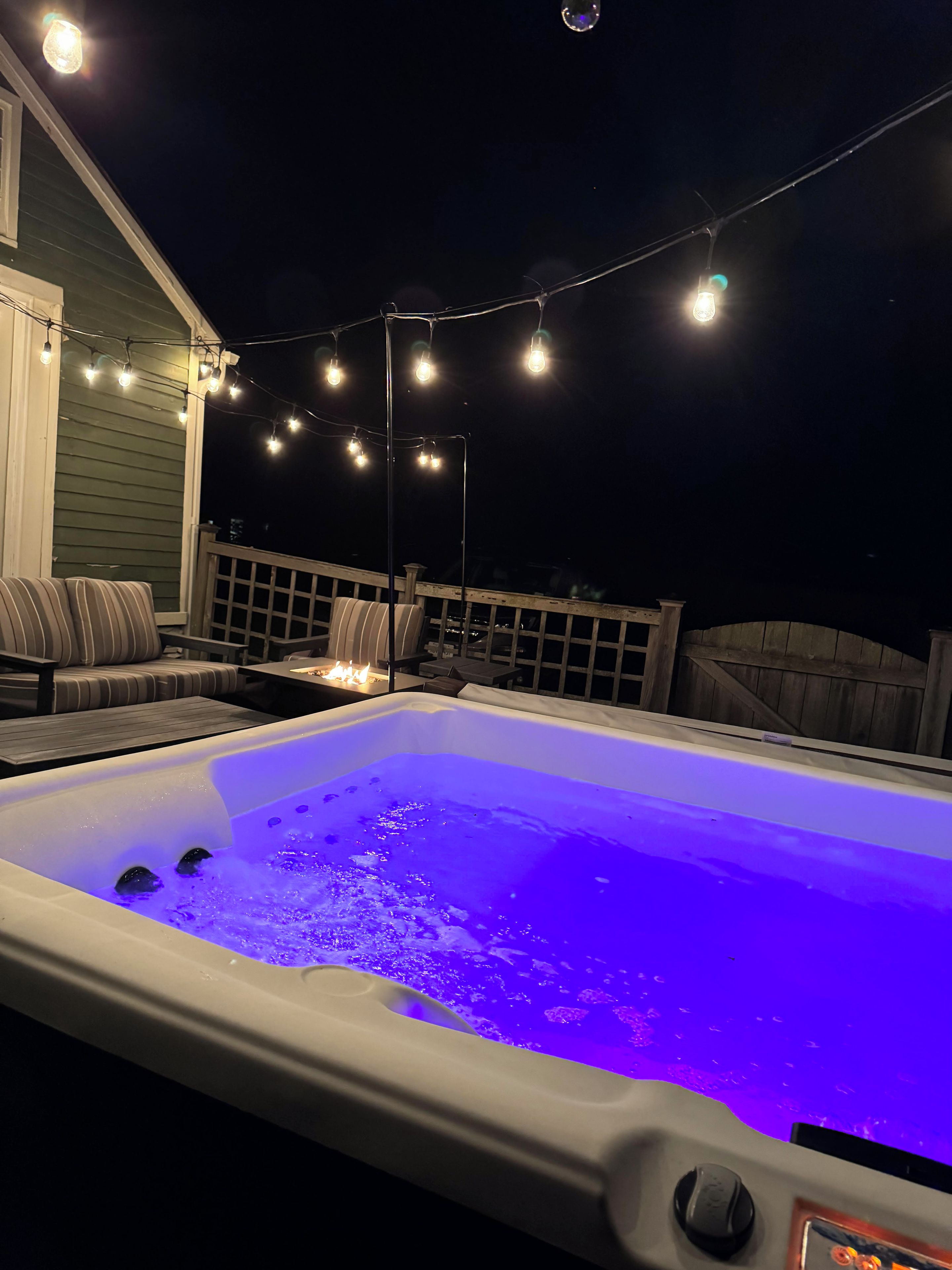 Un jacuzzi con luz azul, rodeado de mobiliario exterior y luces colgantes.