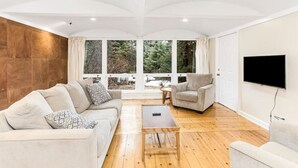 Een moderne woonkamer met een bank, fauteuil en salontafel, grote ramen met uitzicht op bomen, en een flatscreen televisie aan de muur.