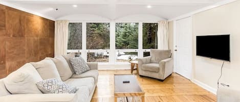 A modern living room with a sofa, armchair, and coffee table, large windows with a view of trees, and a flat-screen TV mounted on the wall.