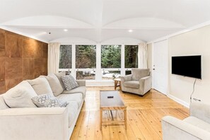 Una sala de estar moderna con sofá, sillón y mesa de centro, ventanas amplias con vistas a árboles y un televisor de pantalla plana montado en la pared.