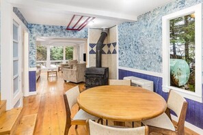 Un comedor con una mesa redonda de madera y sillas, un estufa de leña negra y vistas al exterior a través de una ventana.