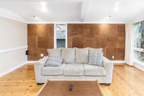 Una sala de estar moderna con un sofá de color claro, cojines estampados, una mesa de centro de madera y un ventanal con cortinas.