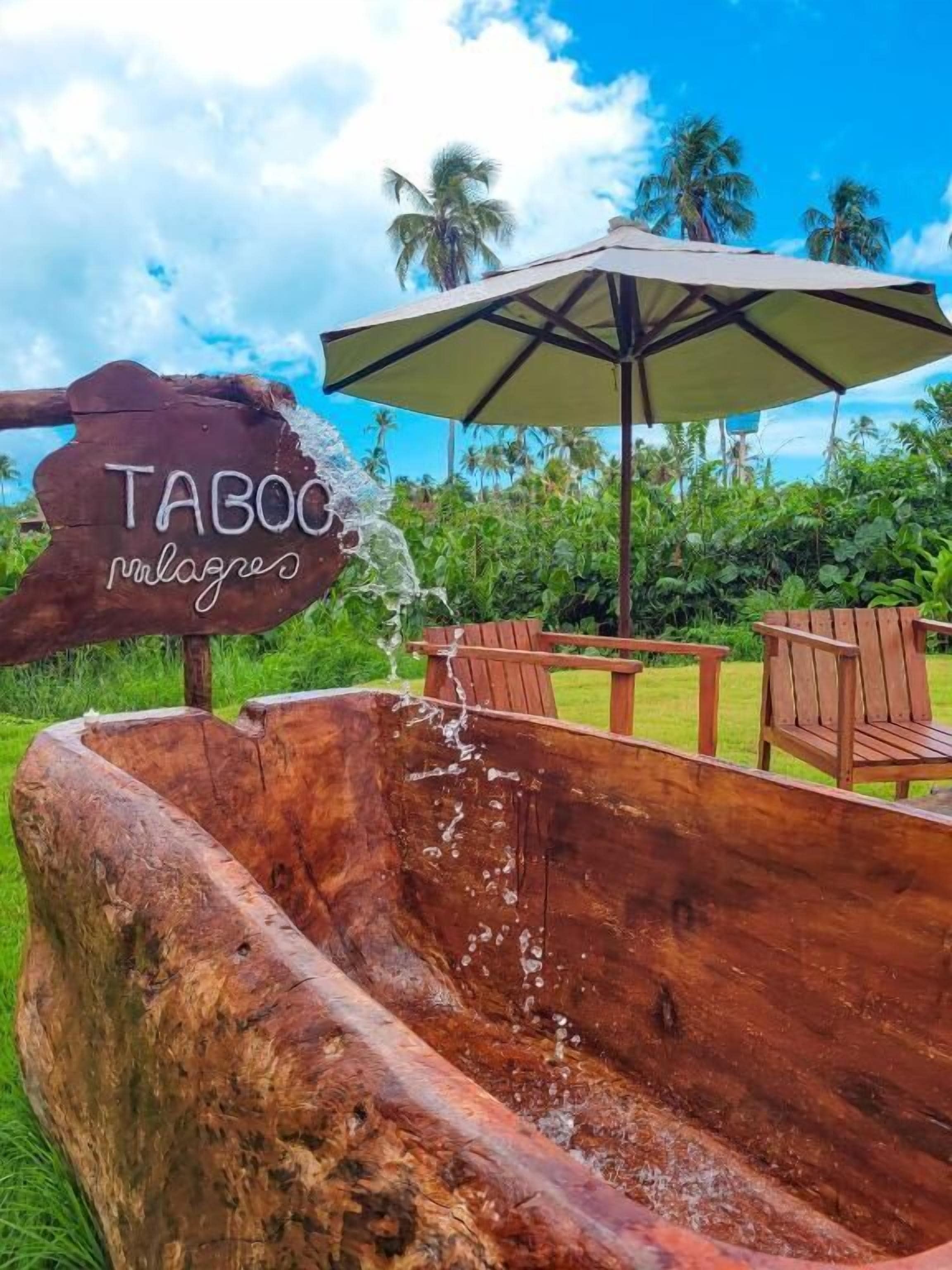 Una bañera de madera con agua brotando, sillas de madera y un letrero que dice "TABOC palaces" bajo un paraguas grande.
