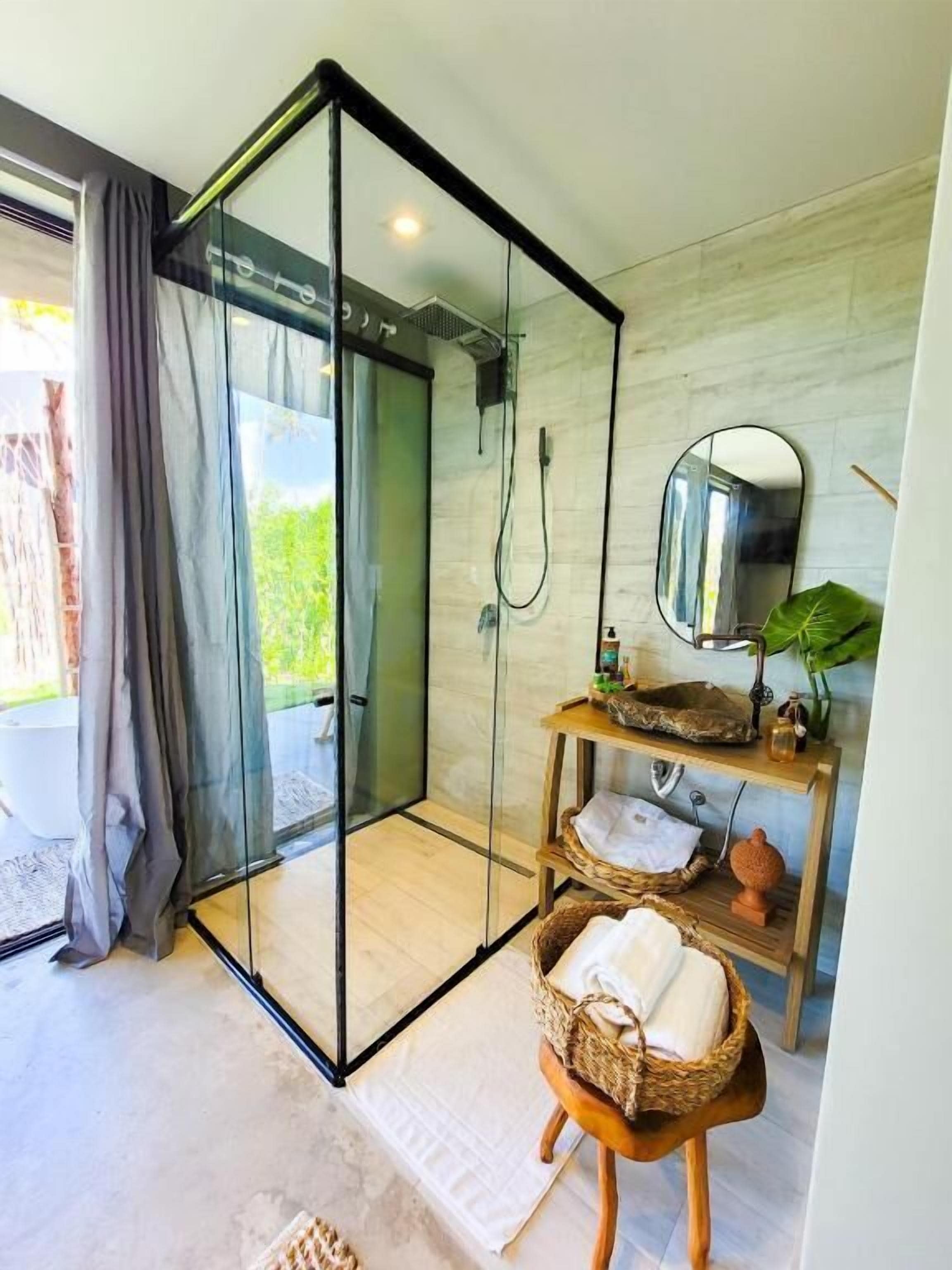 Une salle de bain moderne avec une douche à parois de verre, un meuble-lavabo en bois doté d’un miroir et une vue sur la verdure environnante.
