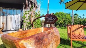 A wooden bowl-shaped planter with a sign reading "TABOO Malagas" in a tropical setting with palm trees and a modern building in the background. - Taboo Milagres (São Miguel dos Milagres)