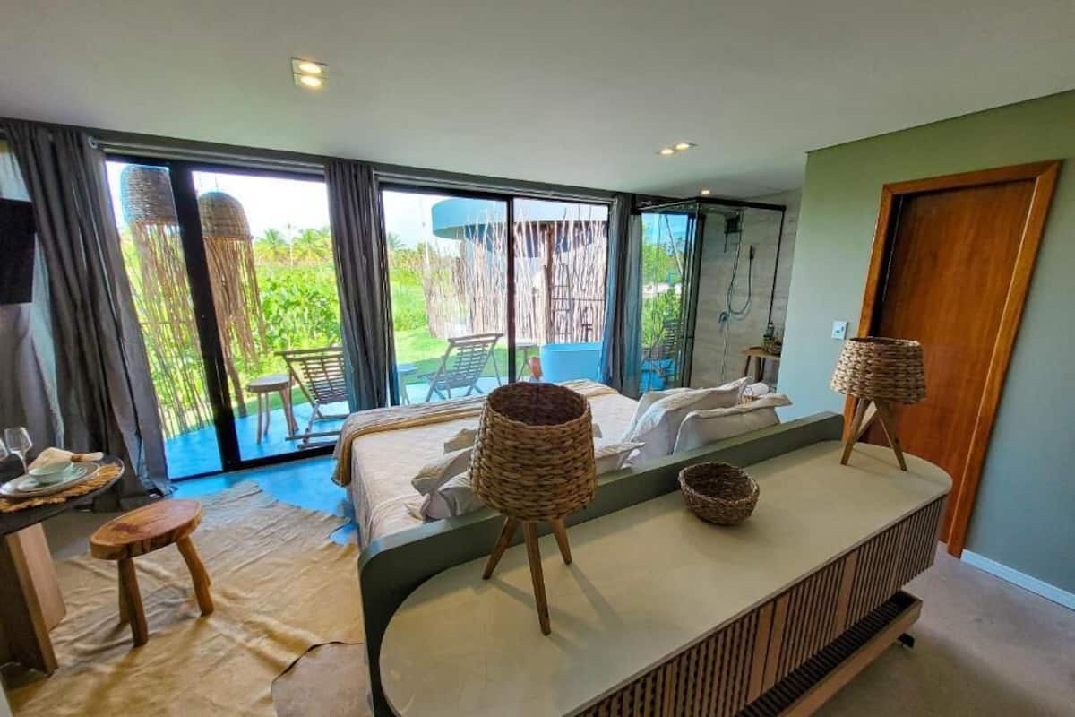 Een moderne slaapkamer met een grote schuifpui naar een balkon, een bed met witte lakens, een klein houten krukje en twee gevlochten manden op een console tafel.