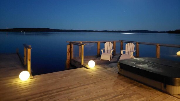 Een houten terras met Adirondack-stoelen en een jacuzzi, met uitzicht op een rustig wateroppervlak in de nacht.