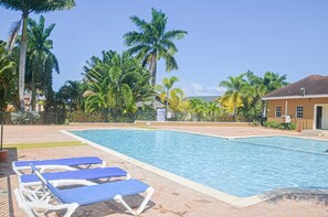 A swimming pool with blue lounge chairs, surrounded by palm trees and a building with a tiled roof. - HAPPY HOLIDAYS at Drax Hall Country Club (Ocho Rios)