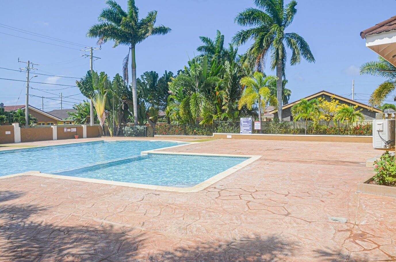 A swimming pool with two sections, surrounded by palm trees and a paved area with a fence.
