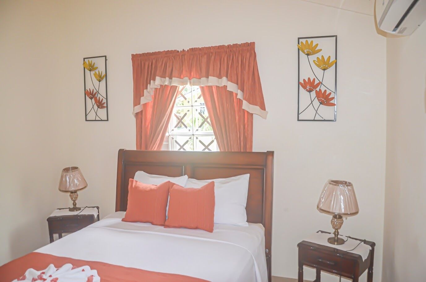 A bedroom with a wooden bed, two bedside tables with lamps, a window with curtains, and floral artwork on the walls.