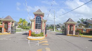 Une entrée fermée par un portail donne accès à un quartier résidentiel, où se trouve un bâtiment central doté d’une grande fenêtre et d’un toit rouge.