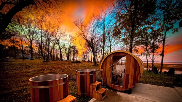 Een houten sauna met een bank en twee grote vaten, op de achtergrond herfstbomen en een zonsondergang.