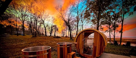 A wooden sauna with a bench and two large barrels, set against a backdrop of autumn trees and a sunset.