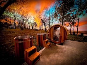 Una sauna de madera con un banco y dos barriles grandes, frente a un fondo de árboles otoñales y un atardecer.