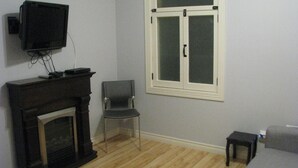 A room with a fireplace, a television mounted on the wall, a window with a white frame, a chair, and a small table. - Budget loft with two beds + sofa bed (Le Citadin) (Rouyn-Noranda)