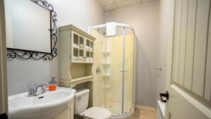 A bathroom with a white sink, toilet, and shower. There is a cabinet with glass doors above the sink. - Loft Economique in the heart of Rouyn-Noranda (Le Citadin) (Rouyn-Noranda)