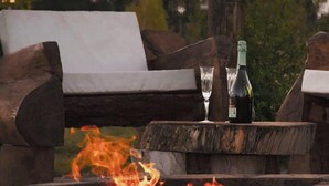 A wooden bench with a bottle and glasses on a table, a fire pit in the foreground, and a tree in the background. - Viento Andino (San Rafael)