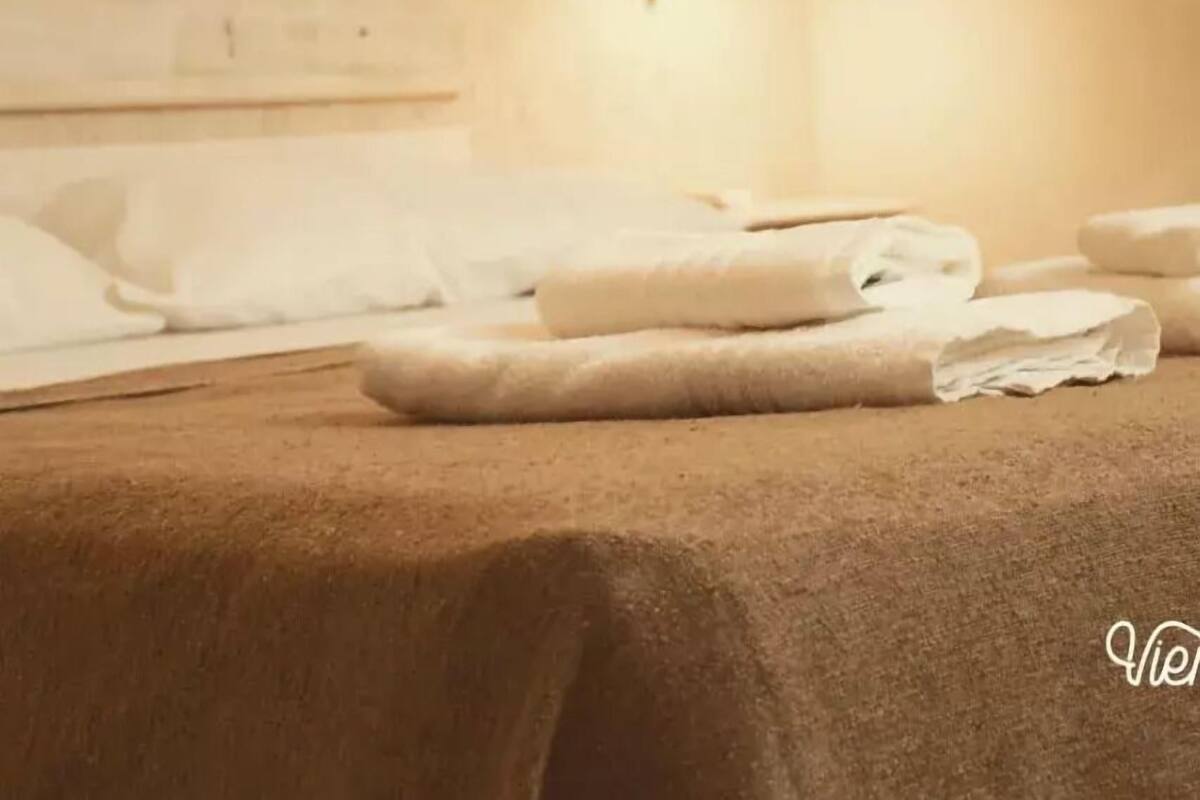 A neatly made bed with white linens and a light brown bedspread, a wall-mounted light fixture, and a partially visible headboard.