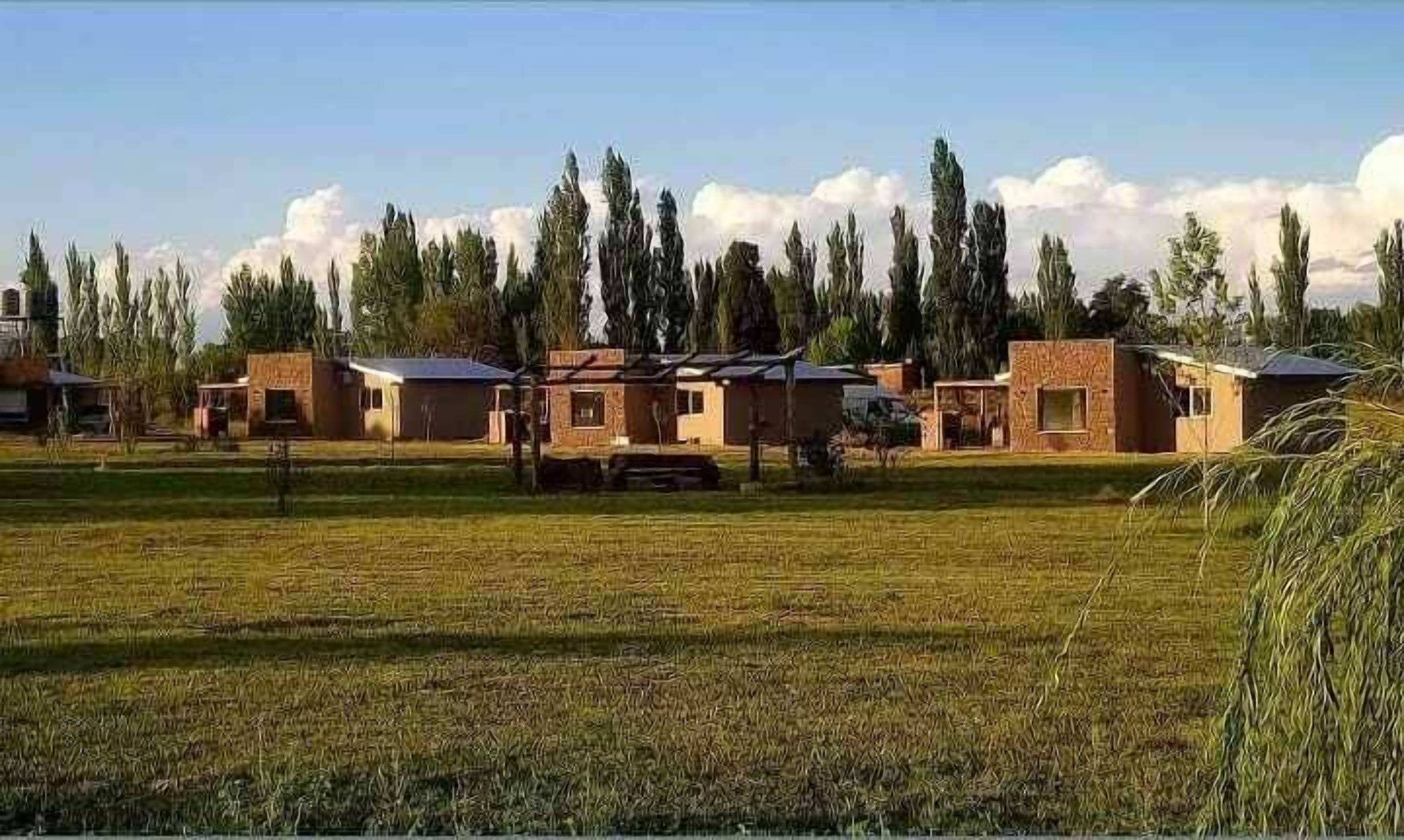 A row of simple, single-story buildings with flat roofs, surrounded by a grassy field and tall trees.