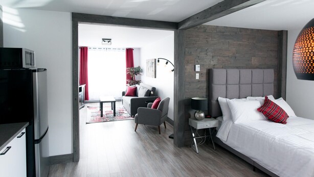A modern hotel room with a bed, bedside table, and a small desk. There is a view of the living area with a sofa and a dining table. - Loft with bed and sofabed in the city centre (Le Court-toit) (Rouyn-Noranda)