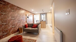 Una sala de estar moderna con pared de ladrillo, un sofá, un reposapiés rojo y una cocina al fondo.