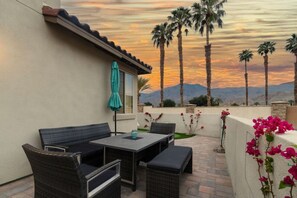 Un patio exterior con mesa y sillas, un paraguas color turquesa, y vistas a palmeras y montañas.