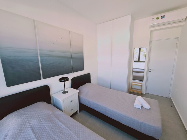 A hotel room with two beds, a nightstand, a large window with a view of the sea, a wardrobe, and a door with a mirror.