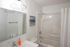 A bathroom with a white sink, a toilet, a bathtub, and a shower. There is a painting on the wall and a geometric light fixture above the mirror. - Suite with cloesed bedroom and sofa bed in the city center (Le Court-toit) (Rouyn-Noranda)