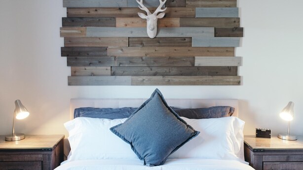 Un dormitorio con cabecera de madera, cabeza de venado montada, cama con ropa de cama blanca y gris, y lámparas de noche.