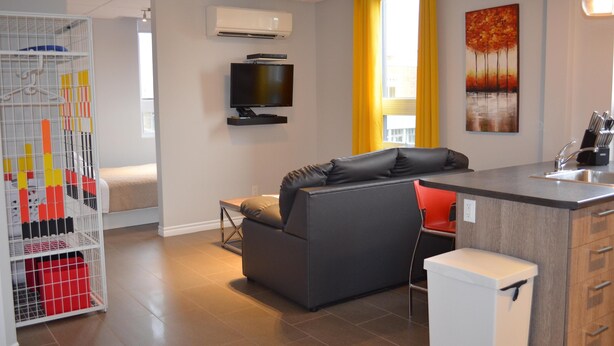 A modern living room with a grey sofa, a small kitchen area, a white storage unit, and a TV mounted on the wall. - Suite with closed bedroom and sofa bed on the rue Principale (Le Chic-urbain) (Rouyn-Noranda)