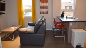 A compact living space with a sofa, a small dining table, a kitchen area with a sink, and a window with yellow curtains. - Suite with closed bedroom and sofa bed on the rue Principale (Le Chic-urbain) (Rouyn-Noranda)