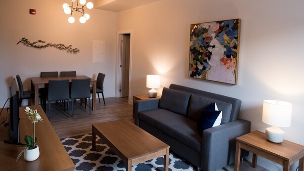 En moderne stue med sofa, sofabord og et spiseområde med stole. Et billede hænger på væggen, og der er en lysekrone.