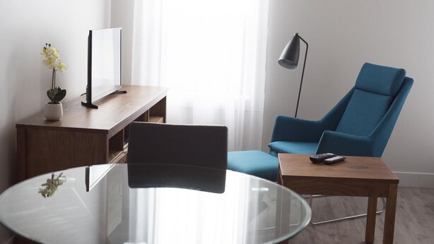 Ein modernes Zimmer mit Glastisch, blauem Sessel, Holzbüro, Blumentopf und Stehlampe.