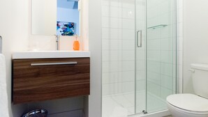 A modern bathroom with a glass shower enclosure, a white toilet, and a wooden vanity with a mirror. - Suite with closed bedroom on rue Gamble Ouest (Le Central) (Rouyn-Noranda)