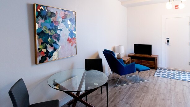 A modern living room with a glass table, blue chairs, a flat-screen TV, and a colorful abstract painting on the wall. - Suite with closed bedroom on rue Gamble Ouest (Le Central) (Rouyn-Noranda)