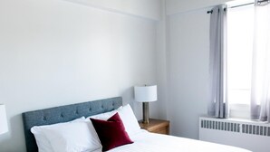 A neatly made bed with white bedding and a red pillow, a bedside table with a lamp, and a window with curtains. - Suite with closed bedroom on rue Gamble Ouest (Le Central) (Rouyn-Noranda)