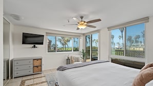 A bedroom with a large bed, a ceiling fan, a TV mounted on the wall, and a view of palm trees through the windows.
