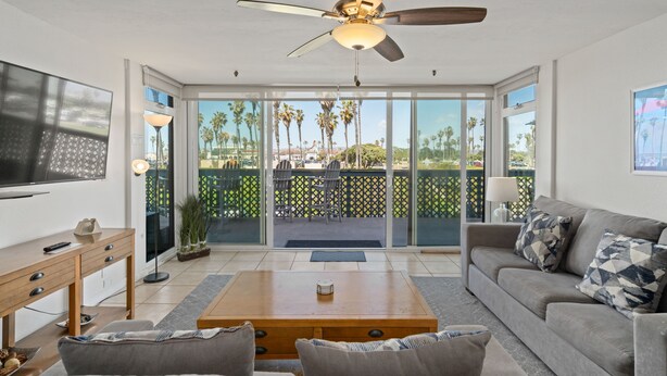 A modern living room with a ceiling fan, a flat-screen TV, a sofa, a coffee table, and a sliding glass door leading to a patio with outdoor furniture and palm trees.