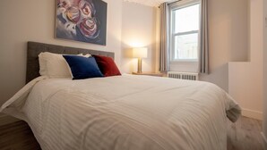 Ein ordentlich bezogenes Bett mit weißer Bettwäsche und bunten Kissen, eine moderne Lampe daneben sowie ein großes abstraktes Gemälde an der Wand.